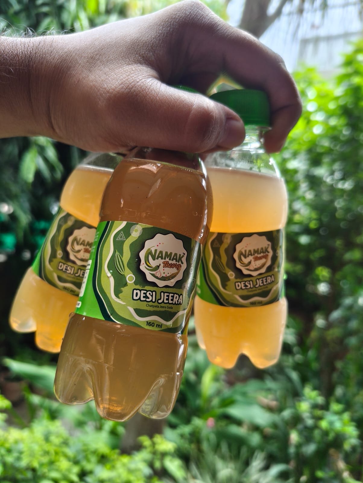 A hand carrying three small Namak Theory Desi Jeera bottles outdoors